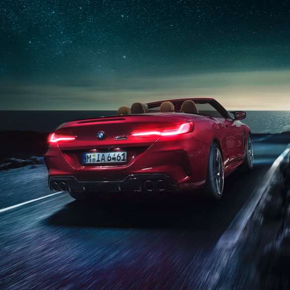 BMW M8 Competition Cabrio, vue de trois quarts arrière. BMW M8 Competition Cabrio en Motegi Rot metallic avec capote ouverte, vue de trois quarts arrière, roulant de nuit sur une route côtière.