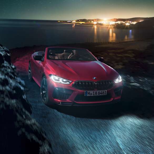 BMW M8 Competition Cabrio, vue de trois quarts face. BMW M8 Competition Cabrio en Motegi Rot metallic avec capote ouverte, vue de trois quarts face, roulant de nuit sur une route côtière.