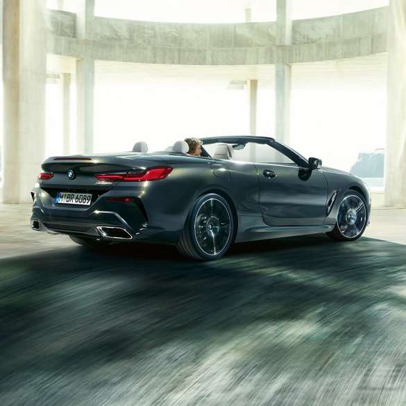BMW M850i xDrive Cabrio, vue de trois quarts arrière. BMW M850i xDrive Cabrio en Dravitgrau metallic avec capote ouverte, vue de trois quarts arrière, en marche devant des immeubles en béton.