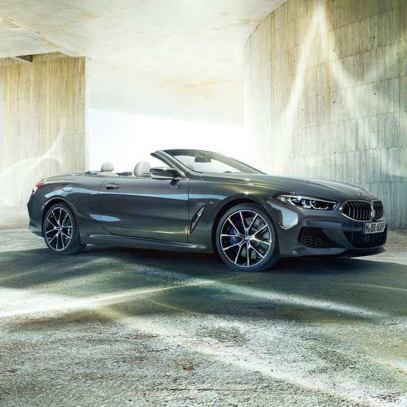 BMW M850i xDrive Cabrio, vue de trois quarts de profil. BMW M850i xDrive Cabrio en Dravitgrau metallic avec capote ouverte, vue de trois quarts de profil, à l’arrêt devant un immeuble en béton.