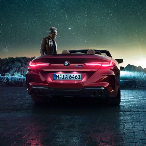 BMW M8 Competition Cabrio, vue arrière. BMW M8 Competition Cabrio en Motegi Rot metallic avec capote ouverte, vue arrière, à l’arrêt avec homme sous un ciel étoilé.