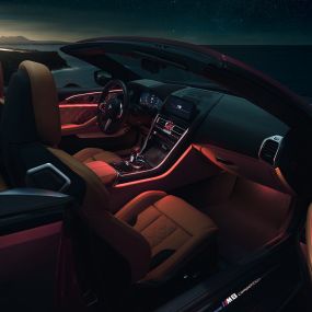 BMW M8 Competition Cabrio, habitacle. BMW M8 Competition Cabrio avec capote ouverte, prise de vue nocturne de l’habitacle avec éclairage d’ambiance rouge.