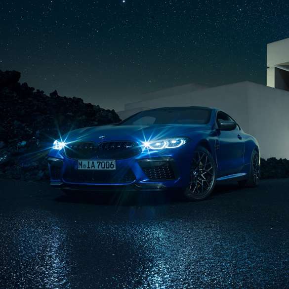 BMW M8 Competition Coupé, vue de trois quarts avant. BMW M8 Competition Coupé en Marina Bay Blau metallic avec phares LED allumés, vue de trois quarts avant à l’arrêt devant une villa, la nuit.