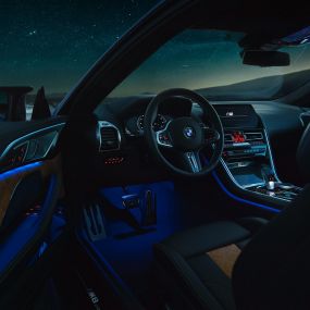Intérieur de la BMW M8 Competition Coupé. BMW M8 Competition Coupé, habitacle vu de nuit, avec volant M en cuir et éclairage d’ambiance bleu, porte du conducteur ouverte.
