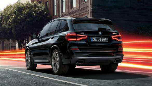 BMW X3 M40i, vue de trois quarts arrière. Extérieur de la BMW X3 M40i en Saphierschwarz metallic, en mouvement, vue de trois quarts arrière devant un paysage urbain avec bandes lumineuses.