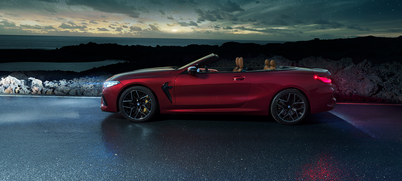 BMW M8 Competition Cabrio, vue de profil. BMW M8 Competition Cabrio en Motegi Rot metallic avec capote ouverte, vue de profil, à l’arrêt sur une route littorale sous un ciel étoilé.