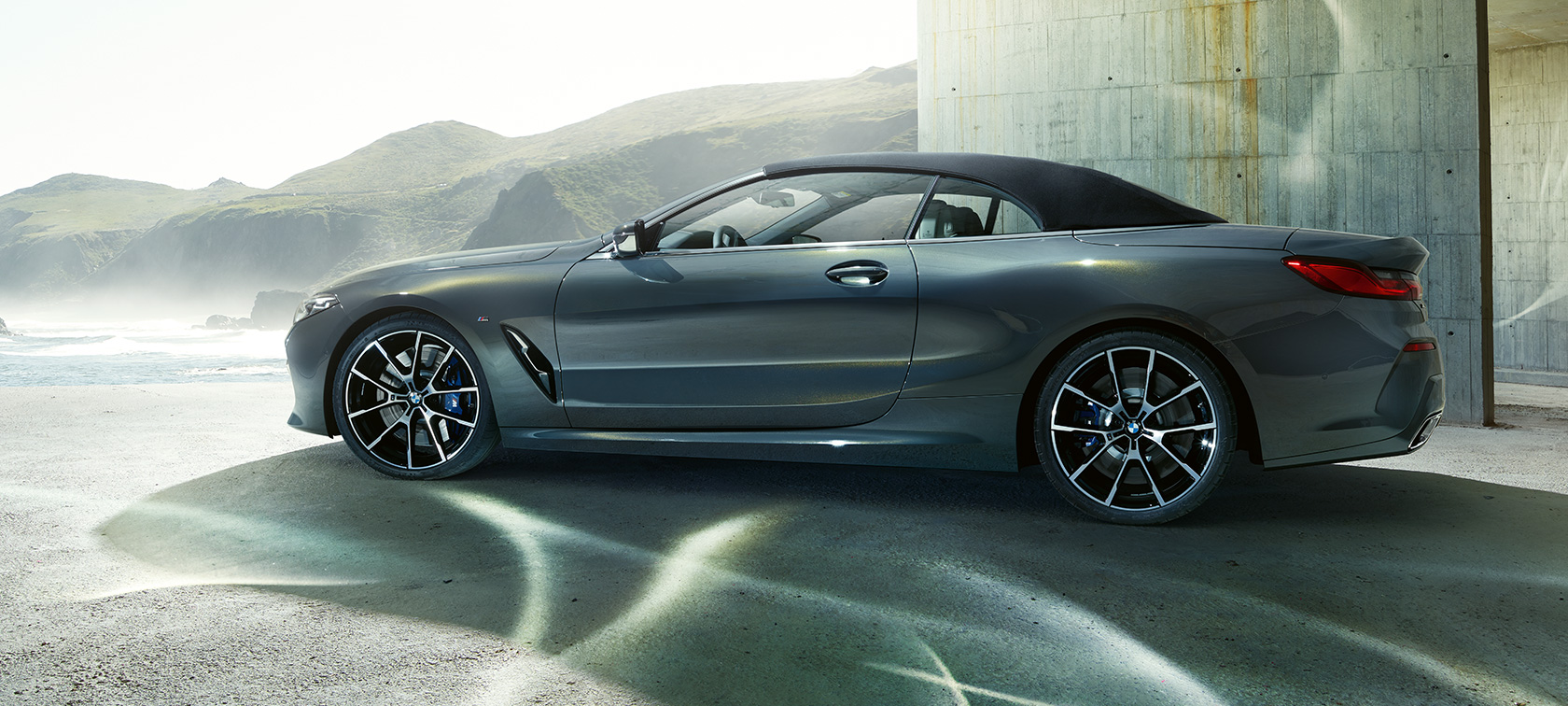 BMW M850i xDrive Cabrio, vue de profil. BMW M850i xDrive Cabrio en Dravit Grau metallic avec capote fermée, vue de profil devant des collines.