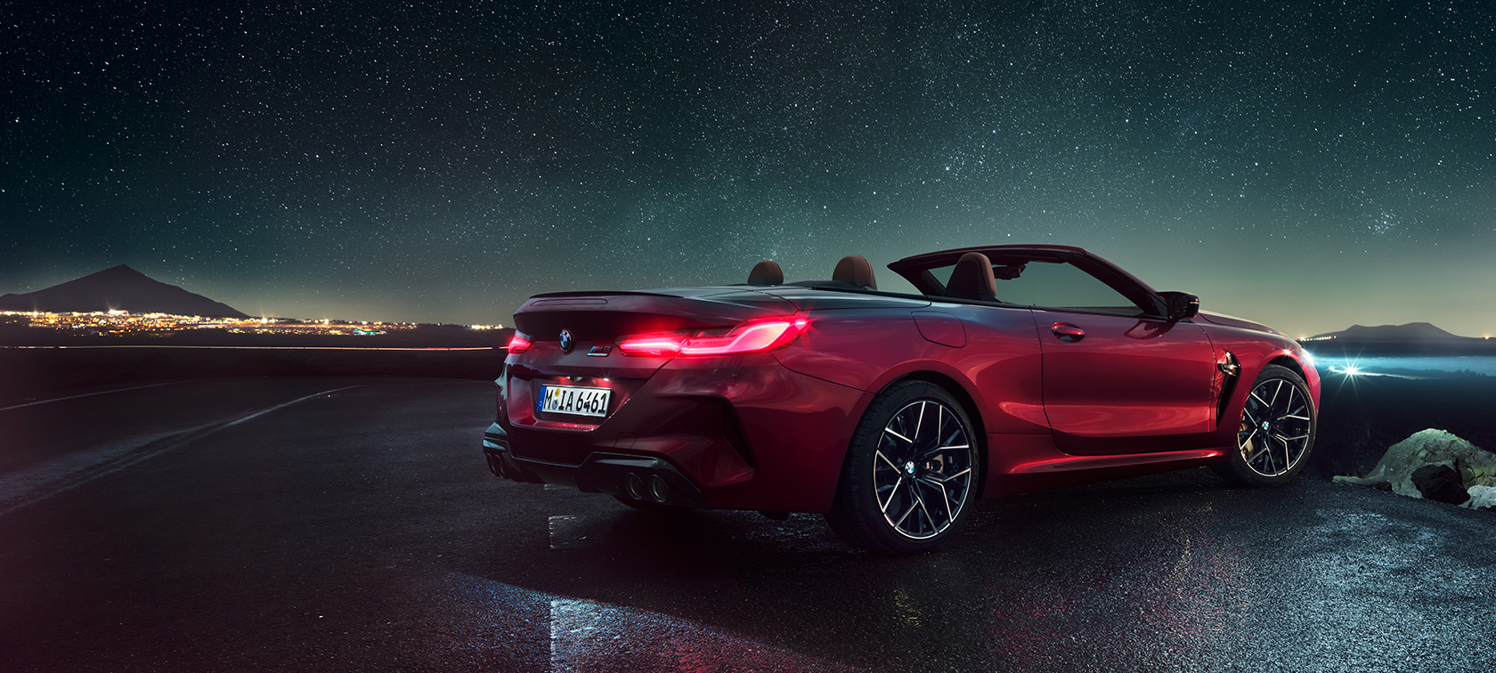 BMW M8 Competition Cabrio, vue de trois quarts arrière. BMW M8 Competition Cabrio en Motegi Rot metallic avec capote ouverte, vue de trois quarts arrière, à l’arrêt sous un ciel étoilé.