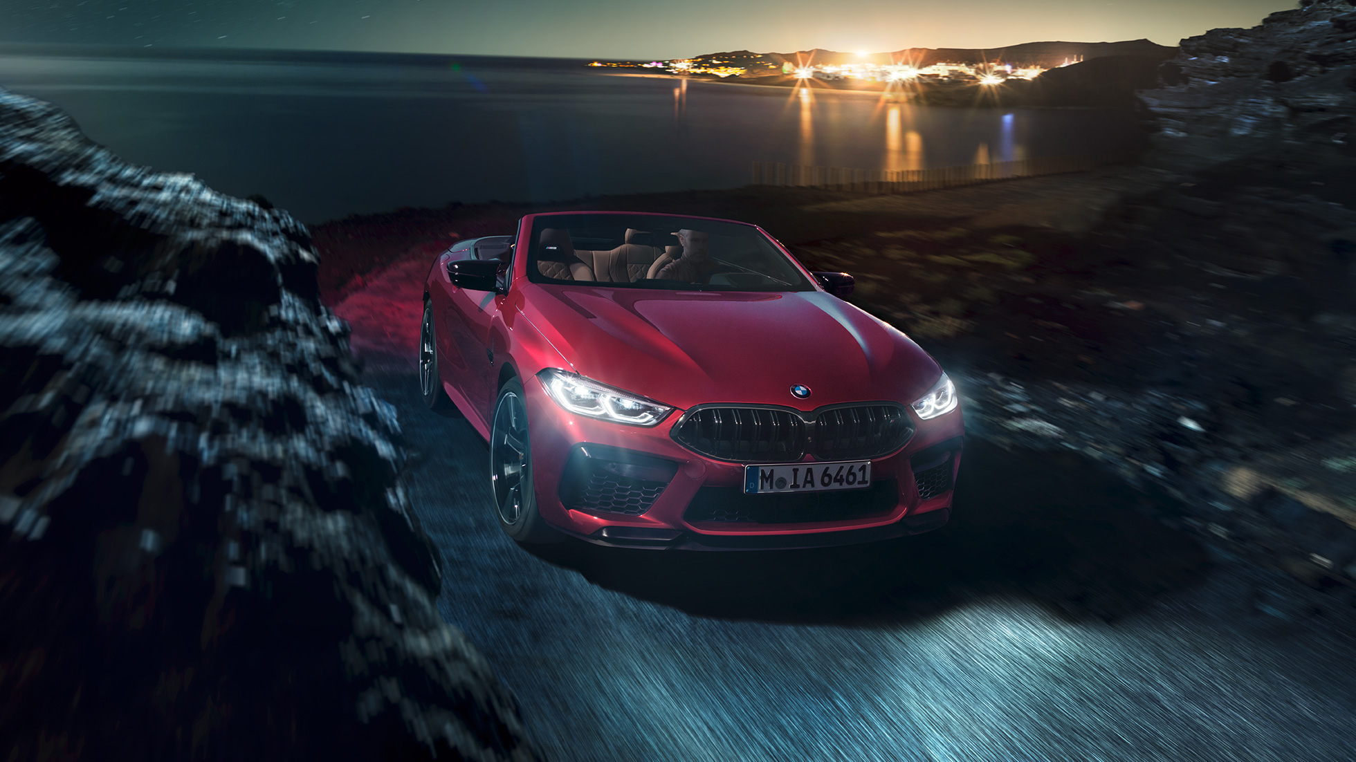 BMW M8 Competition Cabrio, vue de trois quarts face. BMW M8 Competition Cabrio en Motegi Rot metallic avec capote ouverte, vue de trois quarts face, roulant de nuit sur une route côtière.