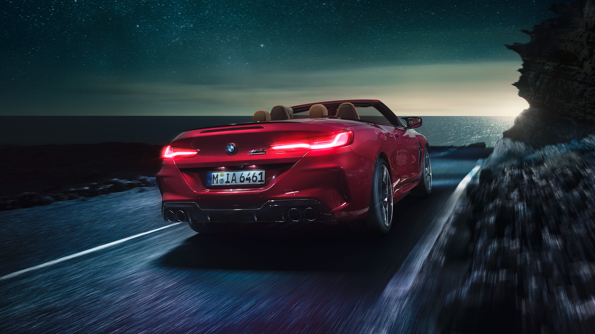 BMW M8 Competition Cabrio, vue de trois quarts arrière. BMW M8 Competition Cabrio en Motegi Rot metallic avec capote ouverte, vue de trois quarts arrière, roulant de nuit sur une route côtière.