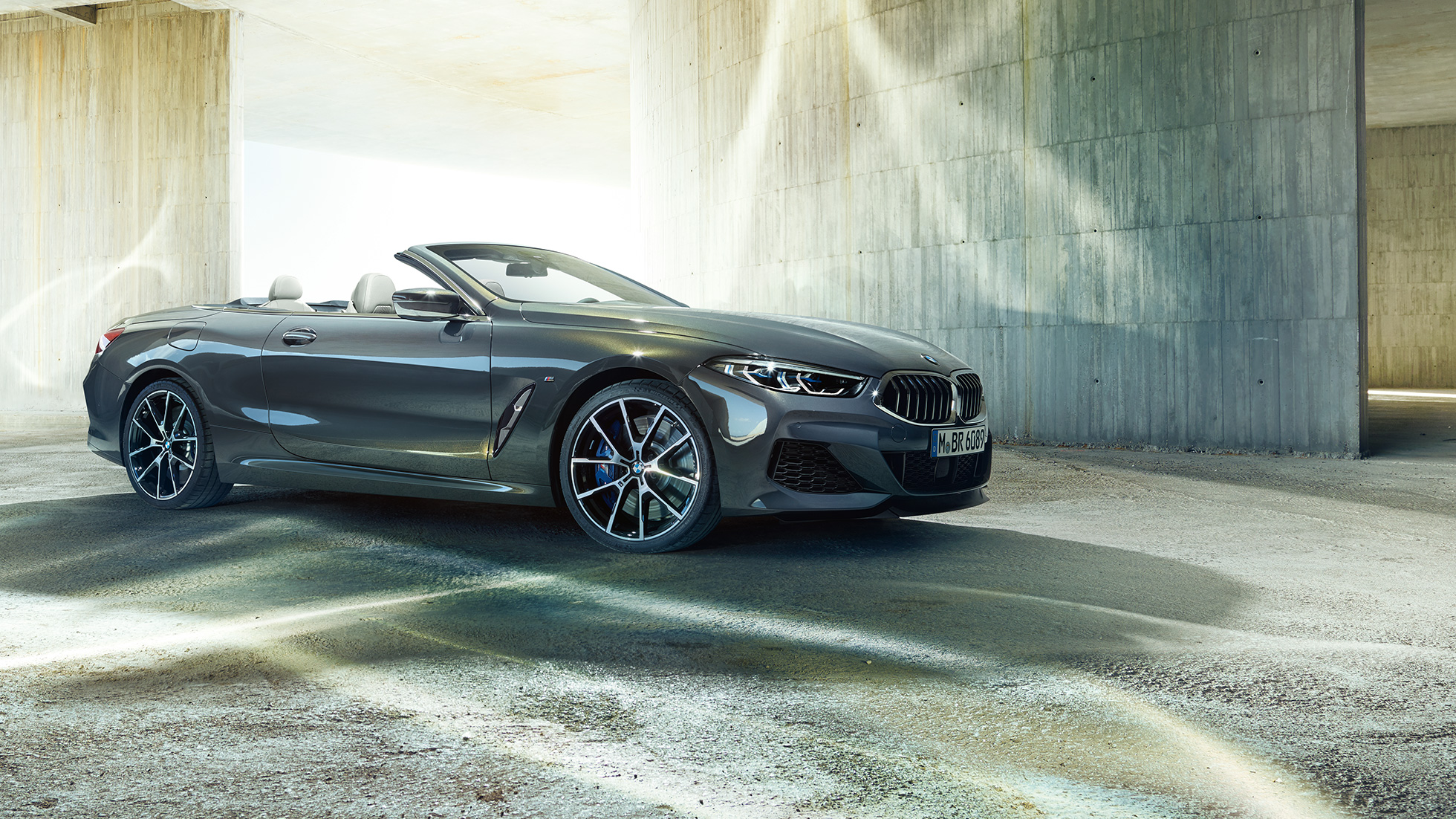 BMW M850i xDrive Cabrio, vue de trois quarts de profil. BMW M850i xDrive Cabrio en Dravitgrau metallic avec capote ouverte, vue de trois quarts de profil, à l’arrêt devant un immeuble en béton.