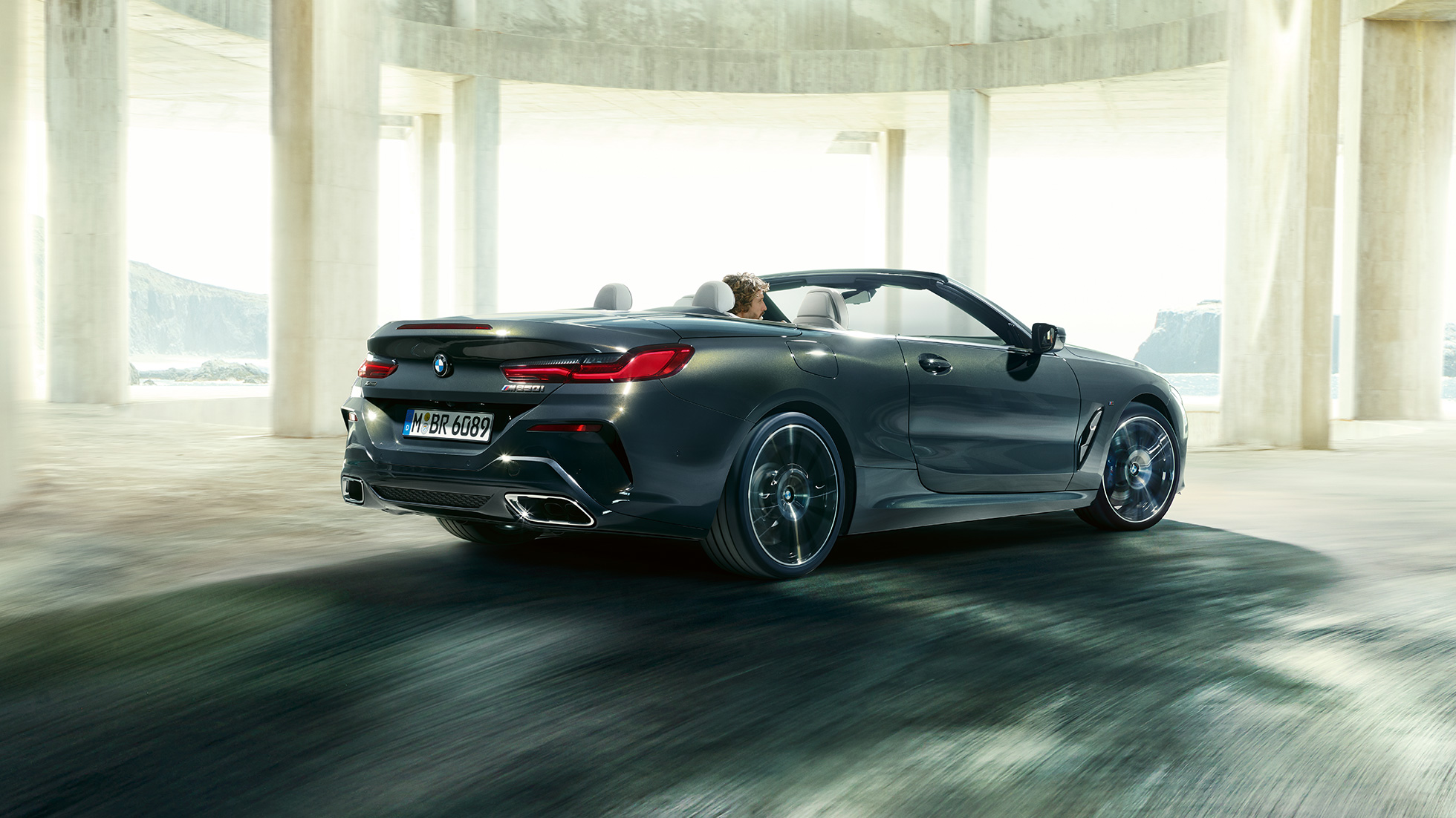 BMW M850i xDrive Cabrio, vue de trois quarts arrière. BMW M850i xDrive Cabrio en Dravitgrau metallic avec capote ouverte, vue de trois quarts arrière, en marche devant des immeubles en béton.