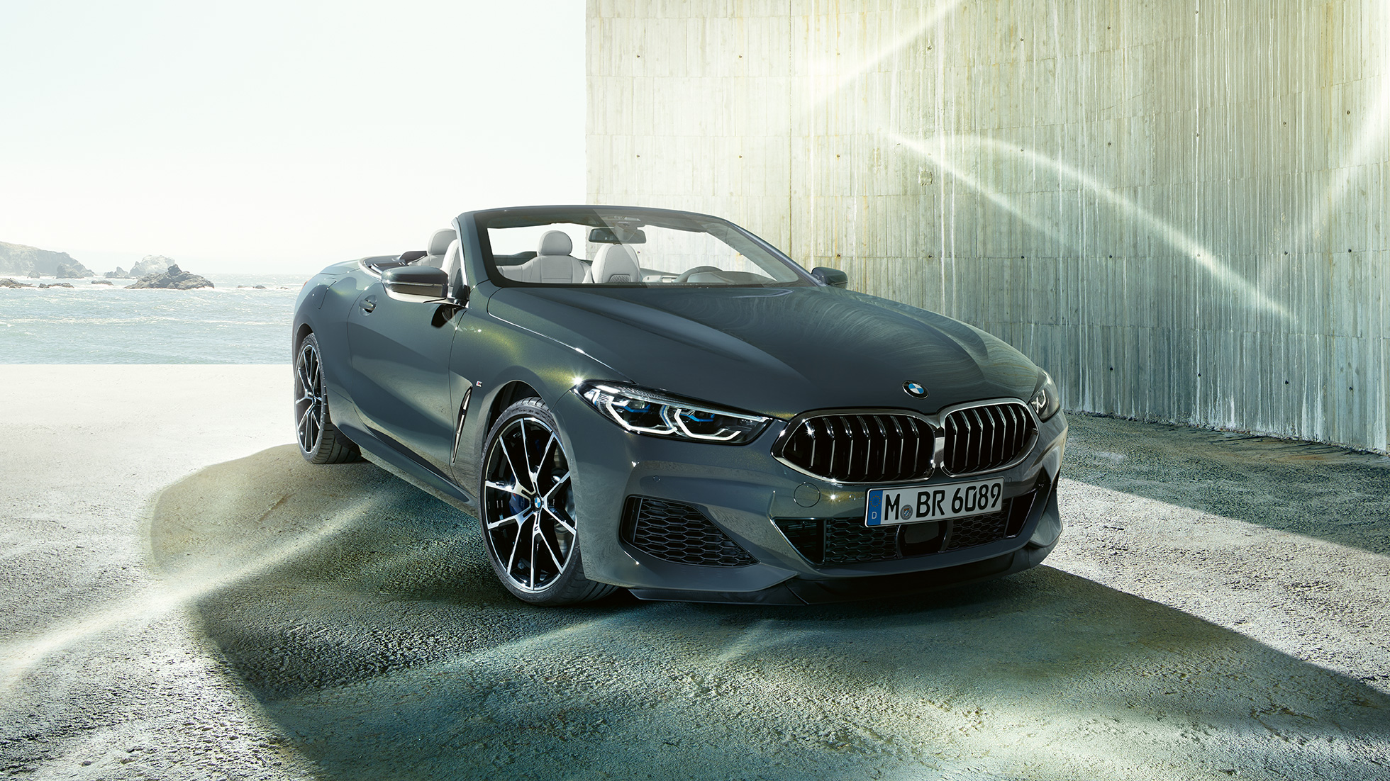 BMW M850i xDrive Cabrio, vue de trois quarts avant. BMW M850i xDrive Cabrio en Dravitgrau metallic avec capote ouverte, vue de trois quarts avant, à l’arrêt devant le littoral.