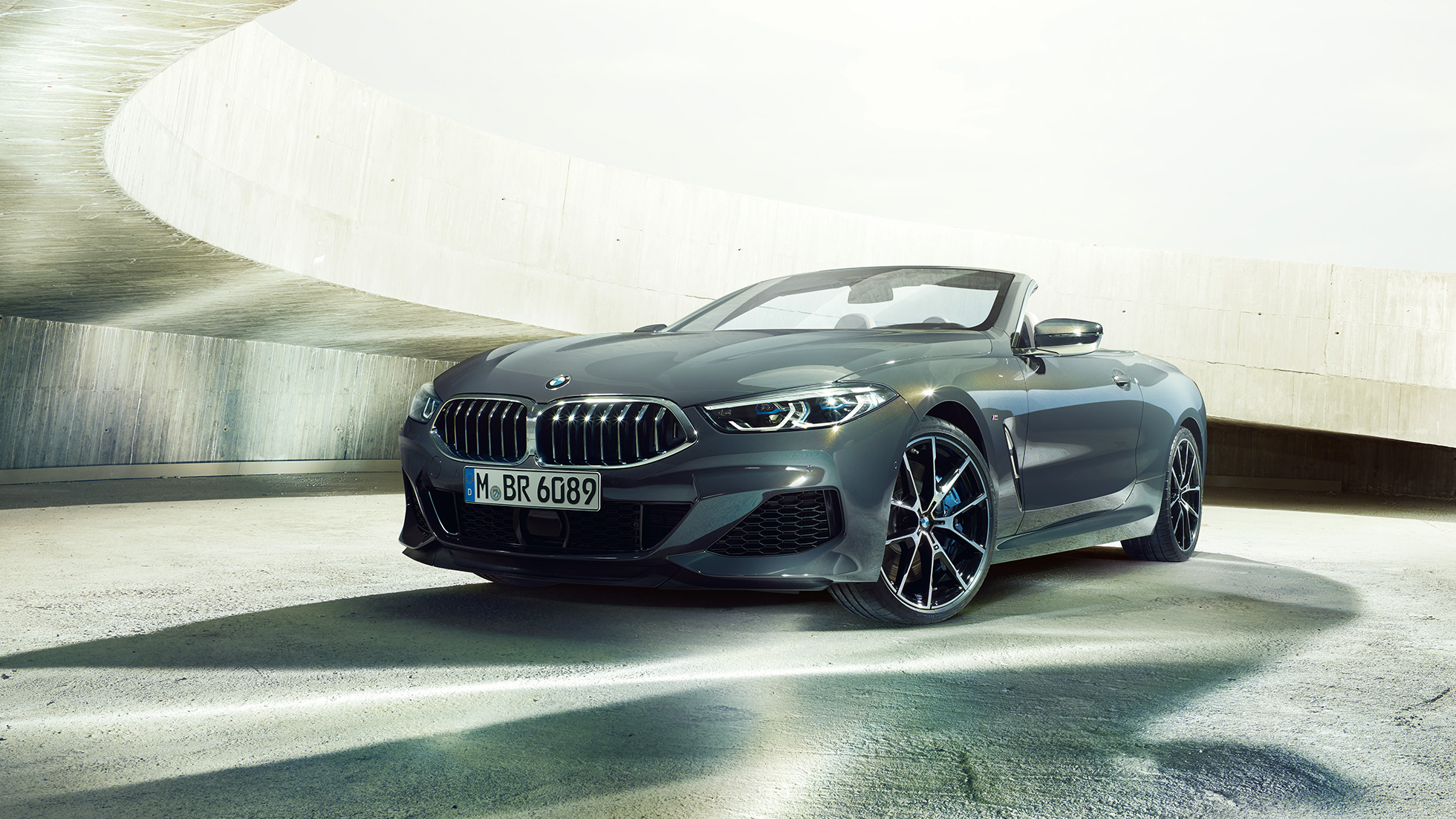 BMW M850i xDrive Cabrio, vue de trois quarts avant. BMW M850i xDrive Cabrio en Dravitgrau metallic avec capote ouverte, vue de trois quarts avant, à l’arrêt dans un bâtiment en béton.
