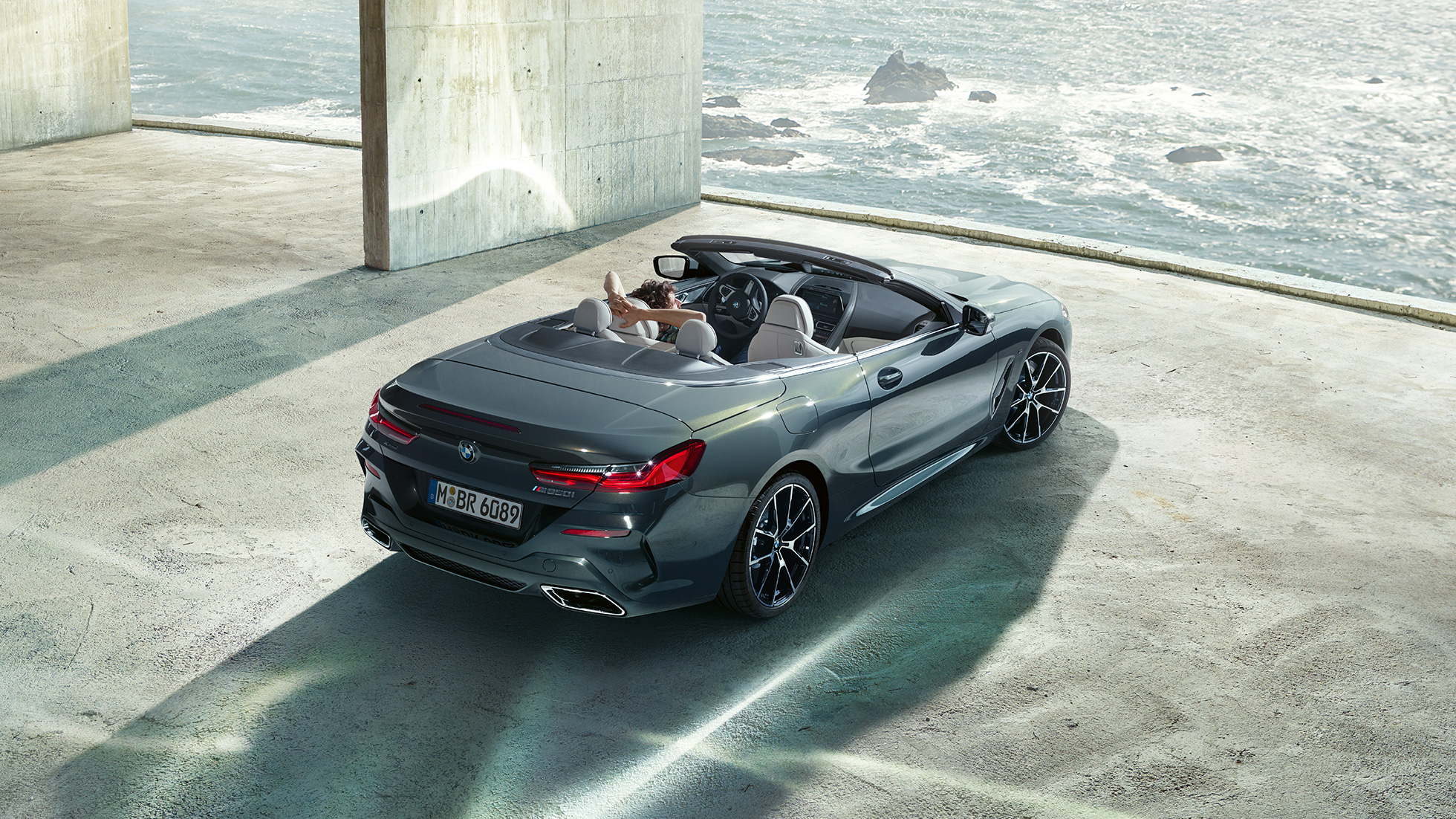 BMW M850i xDrive Cabrio, vue en plongée. BMW M850i xDrive Cabrio en Dravitgrau metallic avec capote ouverte, vue en plongée, en stationnement dans un bâtiment en béton devant l’océan.