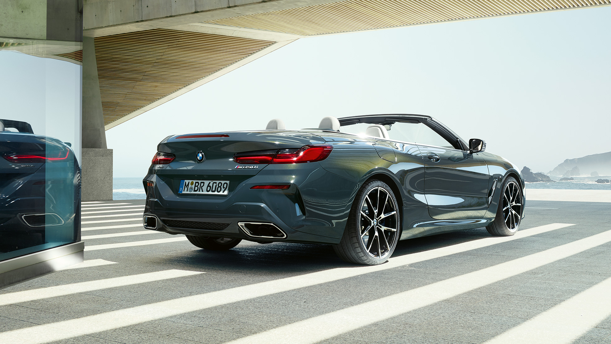 BMW M850i xDrive Cabrio, vue de trois quarts arrière. BMW M850i xDrive Cabrio en Dravitgrau metallic avec capote ouverte, vue de trois quarts arrière, en stationnement avec vue sur l’océan.