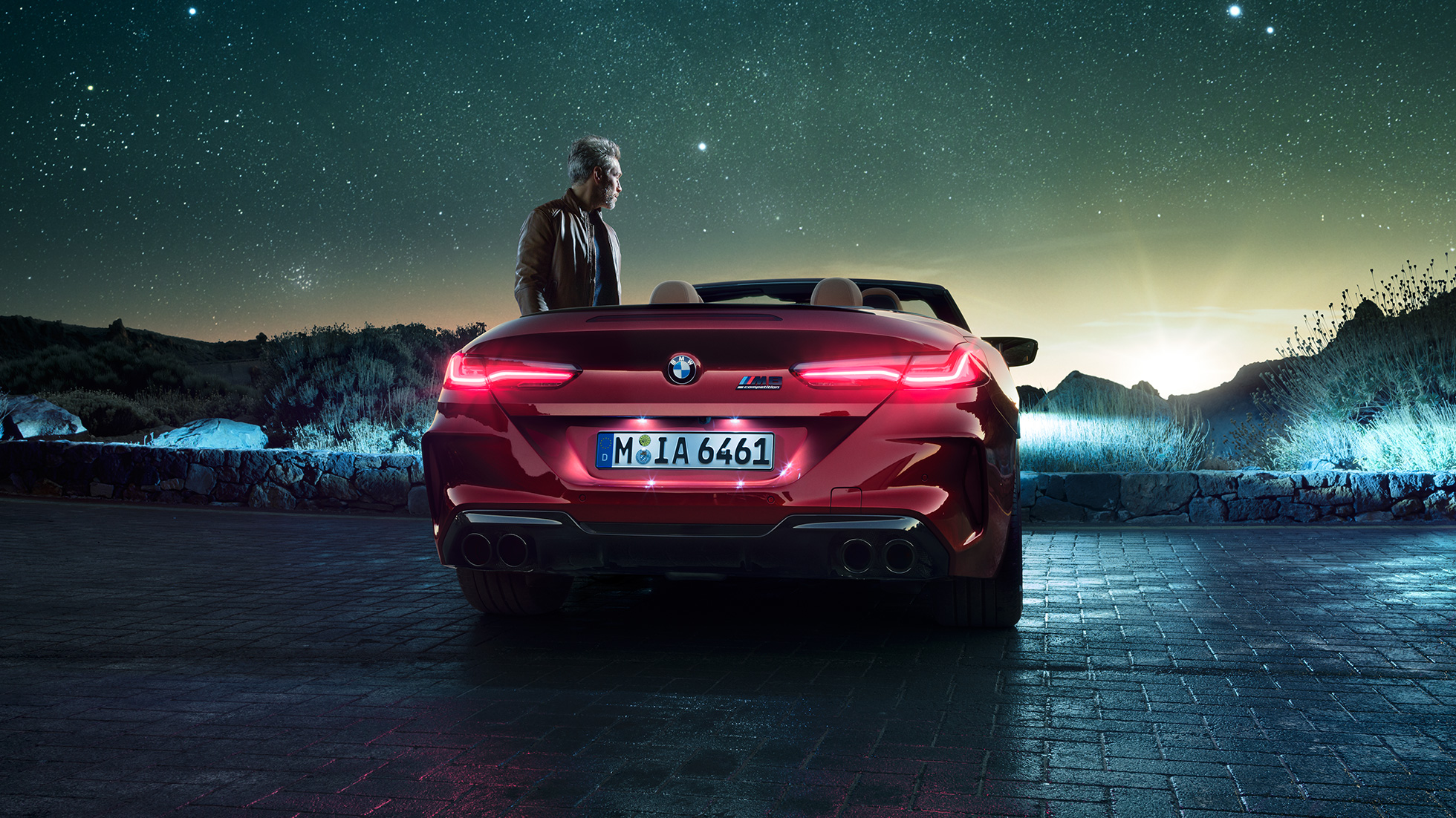 BMW M8 Competition Cabrio, vue arrière. BMW M8 Competition Cabrio en Motegi Rot metallic avec capote ouverte, vue arrière, à l’arrêt avec homme sous un ciel étoilé.