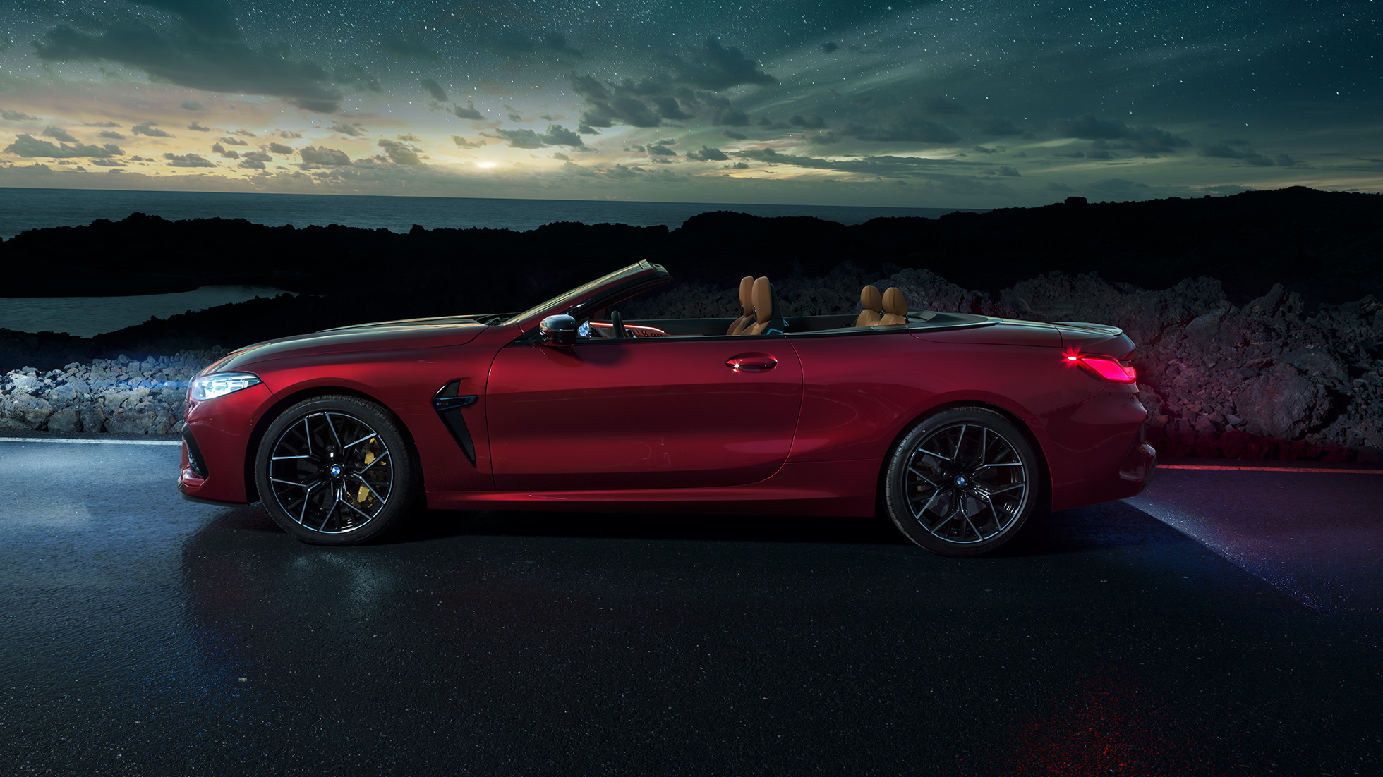 BMW M8 Competition Cabrio, vue de profil. BMW M8 Competition Cabrio en Motegi Rot metallic avec capote ouverte, vue de profil nocturne sous un ciel étoilé.