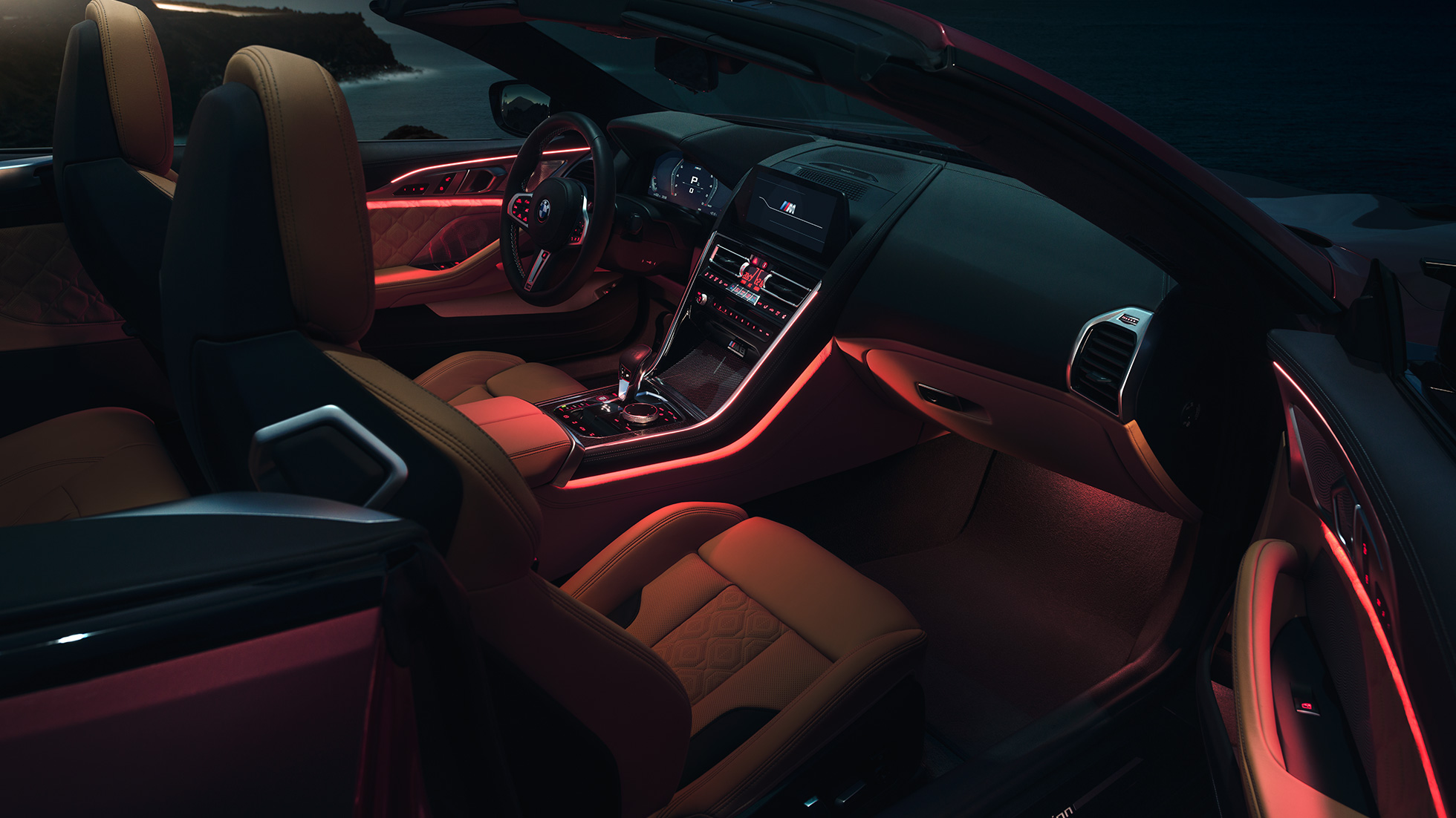 BMW M8 Competition Cabrio, habitacle. BMW M8 Competition Cabrio avec capote ouverte, prise de vue nocturne de l’habitacle avec éclairage d’ambiance rouge.