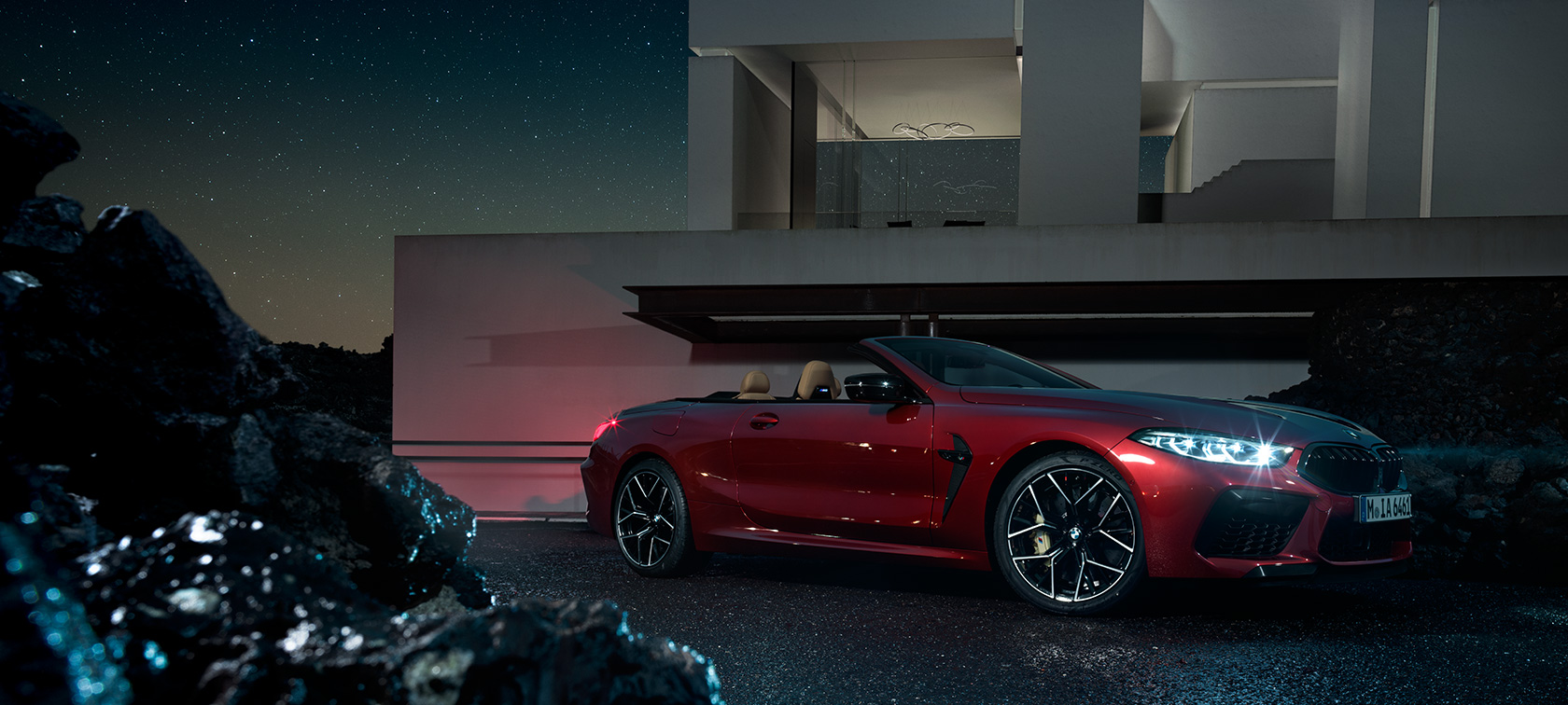 BMW M8 Competition Cabrio, vue de trois quarts de profil. BMW M8 Competition Cabrio en Motegi Rot metallic, vue de trois quarts de profil, à l’arrêt devant une villa, de nuit.