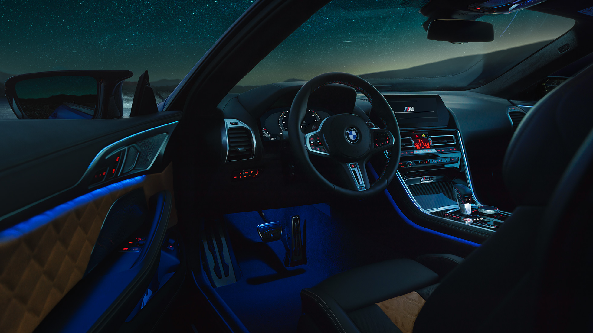 Intérieur de la BMW M8 Competition Coupé. BMW M8 Competition Coupé, habitacle vu de nuit, avec volant M en cuir et éclairage d’ambiance bleu, porte du conducteur ouverte.
